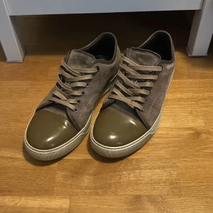 Grå sneakers med olivgröna detaljer från Lanvin - Säljer ett par grå sneakers från Lanvin med olivgröna, blanka tår och matchande snörning. Skorna har en platt sula och är tillverkade i mocka med detaljer i syntet. Perfekta för dig som vill ha en stilren och unik look. Vid några funderingar hörr av dig // William 