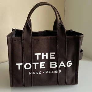 Svart tote bag från Marc Jacobs - Säljer en svart tote bag från Marc Jacobs med tydlig vit text på framsidan. Väskan har två korta handtag och en avtagbar axelrem. Tillverkad i bomullscanvas och rymmer det mesta du behöver till vardagen. Märkets logga syns både på utsidan och på innerfickan.