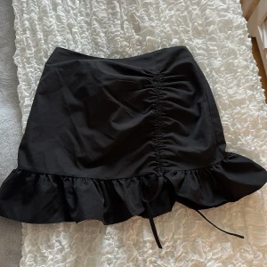 Svart kjol med volang och snörning - Supersnygg svart kjol med volangkant och snörning framtill. Kjolen har en tight passform och är kort i modellen, perfekt för att skapa en trendig look. Enkel att matcha med olika toppar för en cool outfit.