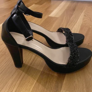 Svarta sandaler med klack och glittrig rem - Snygga svarta sandaler med hög klack och öppen tå. Skorna har en glittrig rem över tårna och justerbart spänne runt vristen. Perfekta för en ute kväll.