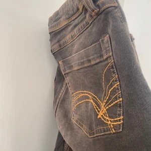 Gråa/bruna  jeansbyxor med orange sömmar - Säljer ett par gråa/bruna bootcut byxor med snygga orange kontrastsömmar. Bakfickan har en unik broderad detalj i orange. Klassisk femficksmodell med knapp och dragkedja framtill. Endast använd 1 gång. 
