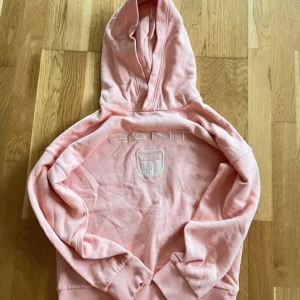 Ljusrosa hoodie från GANT - Säljer en ljusrosa hoodie från GANT med broderad logga och sköld på bröstet. Tröjan har huva och långa ärmar, samt ribbade muddar vid ärmslut och nederkant. Perfekt för en avslappnad stil.