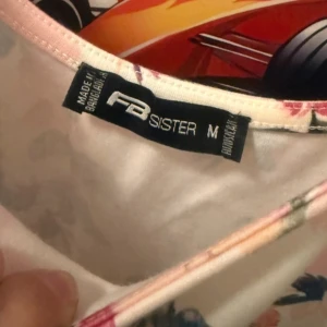 Blommig sommarklänning från Sister - Söt blommig klänning från Sister i vitt med rosa och röda blommor. Klänningen är ärmlös, har rund halsringning och markerad midja. Perfekt för varma dagar och har en lätt, luftig känsla.