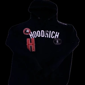 Svart hoodie från Hoodrich med tryck - Säljer en svart hoodie från Hoodrich i storlek S. Tröjan har flera tryck och patchar på bröstet, inklusive ett stort vitt HOODRICH-tryck och röda detaljer. Perfekt för dig som gillar streetwear och vill sticka ut.