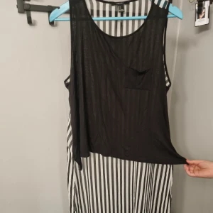 Randig ärmlös klänning med svart overlay - Säljer en ärmlös top från Forever 21 med svart och vit vertikalrandigt mönster och svart transparent overlay med bröstficka. Lätt och luftig modell, perfekt för varma dagar.