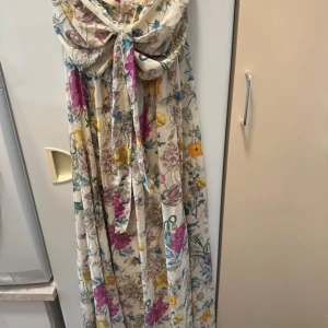 Blommig långklänning med knytband - Säljer en romantisk långklänning med blommigt mönster i färger som rosa, gul, blå och lila på en ljus botten. Klänningen har axelbandslös design, knytband framtill och draperad överdel. Perfekt för sommarens tillfällen.