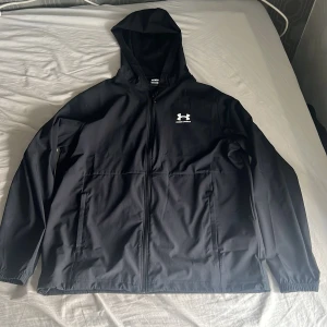 Svart vindjacka från Under Armour - Säljer en svart vindjacka från Under Armour med huva och dragkedja framtill. Jackan har två sidofickor och en diskret logga på bröstet. Perfekt för blåsiga, regniga dagar och enkel att matcha med det mesta. Tröjan har ej använts. Säljer då den är för stor på mig.