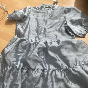 Ljusblå volangklänning med knappar - Säljer en ljusblå klänning med volangdetaljer och knappar framtill. Klänningen har korta puffärmar med resår och hög krage med rynk. Perfekt för dig som gillar romantisk och luftig stil.