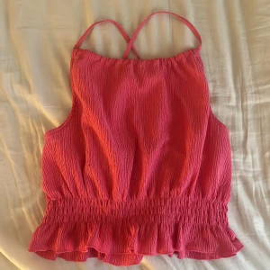Topp - Säljer en söt rosa topp  med smala axelband som korsas i ryggen. Toppen har resår i midjan och en volangkant nedtill för en extra feminin touch. Perfekt för varma dagar!