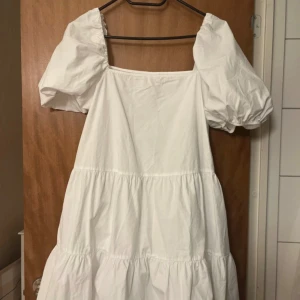 Vit klänning med puffärmar från H&M - Superfin vit klänning från H&M med puffiga kortärmar och fyrkantig ringning. Klänningen har en luftig passform och volanger i kjolen, perfekt för varma dagar. Enkel och stilren design som känns både trendig och klassisk.