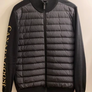 Svart quiltad cardigan från Canada Goose - Säljer en svart quiltad cardigan från Canada Goose med dragkedja och ribbad krage. Cardiganen har långa ärmar och stor logotyp i guld på ena ärmen. Perfekt för dig som vill ha en stilren och sportig look.