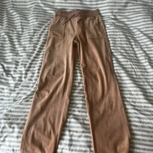 Beige mjukisbyxor från Juicy Couture - Säljer ett par beige mjukisbyxor från Juicy Couture med bred resår i midjan och stora fickor med knappar bak. Byxorna har en avslappnad passform och är gjorda i mjukt material som känns skönt mot huden.