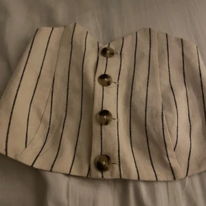 Vit randig tubtopp från Bershka - Säljer en krämvit tubtopp från Bershka med svarta tunna ränder och snygga knappar framtill. Toppen har smockad rygg för extra komfort och sitter fint på kroppen. Perfekt till sommaren och enkel att matcha med jeans eller kjol.