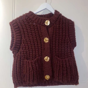  stickad väst med stora guldknappar - Mysig stickad väst med grov struktur och stora, runda guldknappar framtill. Västen har två praktiska fickor och en hög krage. Perfekt för lager-på-lager och en snygg detalj till din outfit.