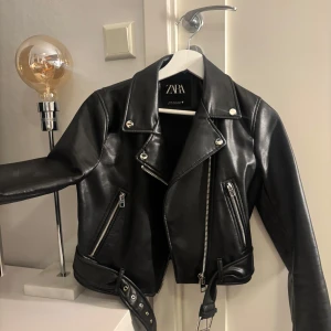 Svart skinnjacka från Zara - Hej!🩷Jag säljer en svart skinnjacka från Zara. Jackan har silvriga dragkedjor, knappar och bälte i midjan. Snygg krage och detaljerade sömmar. Jackan är felfri och i nytt skick, och har endast använts ett fåtal gånger. Pris går att diskutera vid snabb affär.🥰