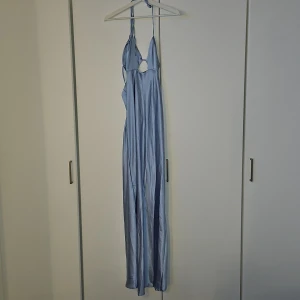 Ljusblå långklänning i satin från Boohoo - Säljer en ljusblå långklänning i satin från Boohoo med smala axelband och knyt i ryggen. Klänningen har en liten cut-out framtill och en elegant, glansig finish. Perfekt för speciella tillfällen när du vill sticka ut.