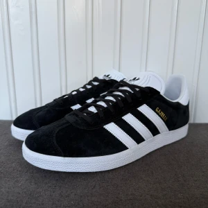 Svarta Adidas Gazelle  - Säljer ett par klassiska Adidas Gazelle sneakers i svart mocka med vita detaljer och de ikoniska tre ränderna på sidan. Skorna har snörning, vit sula och guldtext på sidan. Perfekta för en stilren och sportig look.