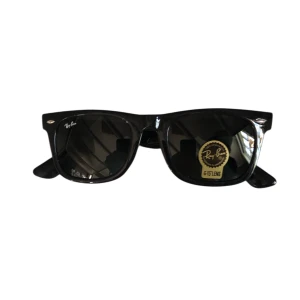 Svarta solglasögon från Ray-Ban - Klassiska svarta solglasögon från Ray-Ban med Wayfarer-form. Glasögonen har svarta bågar i plast och mörka linser. Ray-Ban-logga syns på båda skalmarna och linsen. Levereras med originalfodral och ask.