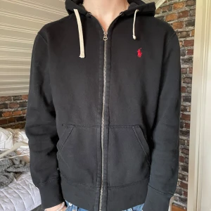 Svart hoodie med dragkedja från Ralph Lauren - Säljer en svart hoodie från Ralph Lauren med dragkedja och huva. Tröjan har två fickor framtill, vita snören i huvan och en liten röd logga broderad på bröstet. Perfekt för en avslappnad stil. Skriv för fler bilder
