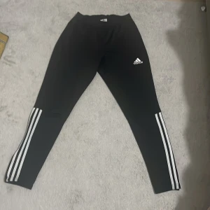 Svarta träningstights från Adidas med vita ränder - Svarta träningstights från Adidas med klassiska vita ränder längs benen och Adidas-logga på låret. Tightsen har en tight passform och är perfekta för träning eller sportiga outfits.