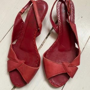 Röda sandaler med öppen tå och rosettdetalj - Säljer ett par röda sandaler med öppen tå och snygg rosettdetalj framtill. Skorna har en smal rem runt hälen och platt sula, perfekta för varma dagar. Klassisk och stilren design som passar till många outfits.