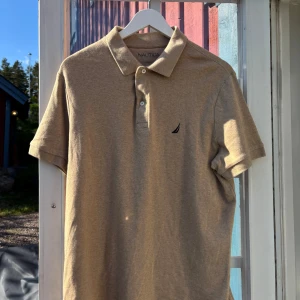 Beige pikétröja från Nautica - Säljer en stilren beige pikétröja från Nautica med klassisk krage och två knappar framtill. Tröjan har korta ärmar och en liten broderad logga på bröstet. Perfekt för en avslappnad och snygg look. Skriv till mig vid frågor eller om du vill ha fler bilder!🙏