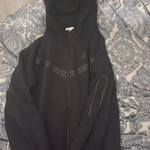 Svart hoodiejacka från Nike - Svart hoodiejacka från Nike med dragkedja framtill och huva. Jackan har diskreta grå detaljer över bröstet och en ficka med dragkedja på ena ärmen. Perfekt för en sportig och avslappnad stil.