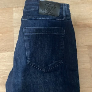 Mörkblå jeansbyxor från SikSilk - Snygga mörkblå jeansbyxor från SikSilk med klassisk femficksdesign och läderpatch med logga bak i midjan. Jeansen har en rak passform och är tillverkade i ett stretchigt denimtyg för extra komfort.