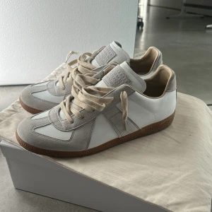 Maison Margiela Replica sneakers i vitt och beige - Maison Margiela Replica sneakers i vitt skinn och beige mocka med klassisk gummisula. Snygg retrostil med rund tå och snörning. Perfekta för dig som gillar minimalistisk och stilren design. Obs! Box finns inte därav priset! Skorna är i topp skick inga defekter eller liknande! 
