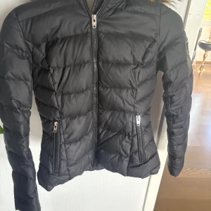 Svart dunjacka med fuskpäls från Rockandblue - Svart pufferjacka från Rockandblue med dragkedja framtill och två dragkedjeförsedda fickor. Jackan har huva med brun päls och Rockandblue-logga på ärmen. Perfekt för kyliga dagar. Storleken är xs/s. Köpte den för 2500 men kom ej till användning.