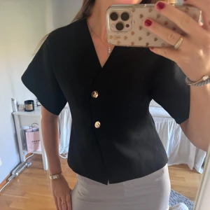 Svart kortärmad kavajkofta med knappar - Snygg svart blus med korta ärmar och två dekorativa guldknappar framtill. Blusen har en v-ringad design och är lätt figursydd för en stilren look.✨