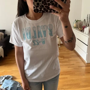 Vit t-shirt med blå text och stenar - Säljer en vit t-shirt med glittrig blå text och siffror framtill. T-shirten har klassisk passform, rund halsringning och korta ärmar. Perfekt för en avslappnad stil och enkel att matcha med jeans eller shorts.