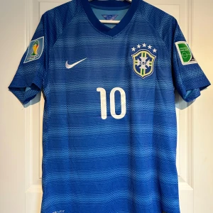 Brasilien 2014 borta tröja med Neymar Jr 10  - Säljer en blå Brasilien fotbollströja med Neymar Jr och nummer 10 på ryggen. Tröjan har korta ärmar, vita detaljer och officiella FIFA World Cup 2014 och Football for Hope patchar på ärmarna. Perfekt för fans av landslaget🇧🇷