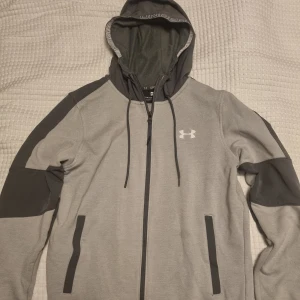 Grå hoodiejacka från Under Armour - Grå hoodiejacka från Under Armour med  dragkedja. Jackan har huva med dragsko, två fickor framtill och logga på bröstet. 
