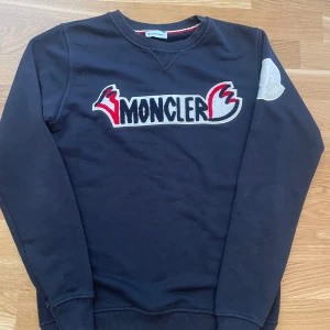 Svart sweatshirt från Moncler med broderad logga - Svart sweatshirt från Moncler med stor broderad logga i vitt, rött och svart framtill. Tröjan har rund halsringning, ribbade muddar och en stor vit Moncler-patch på ena ärmen. Perfekt för en stilren och sportig look.