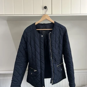 Marinblå quiltad jacka från Soyaconcept - Snygg marinblå quiltad jacka från Soyaconcept i storlek M. Jackan har rund halsringning, dragkedja framtill och två fickor med dragkedja. Perfekt för övergångssäsonger och enkel att matcha med olika outfits.
