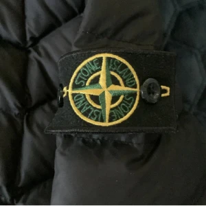 Svart pufferjacka från Stone Island - Säljer en svart pufferjacka från Stone Island med klassisk patch på ärmen. Jackan har lång ärm, dragkedja framtill och två snedställda fickor med dragkedja. Perfekt för kyliga dagar och har en modern, sportig look. OBS jackan har en defekt i fickan, det är ett litet hål där därför är priset lågt