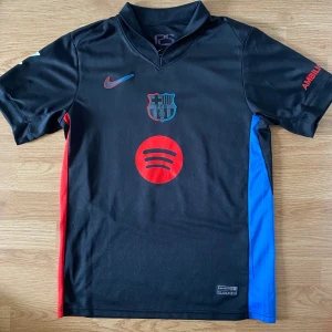 FC Barcelona svart bortatröja, Lamine Yamal 19 - Säljer en svart FC Barcelona bortatröja från Nike med Lamine Yamal 19 på ryggen. Tröjan har röda och blå detaljer på sidorna, klubbmärke och sponsortryck på bröstet samt UNHCR-logga bak. Kortärmad och tillverkad i Dri-FIT-material. För optimal kvalité och feel. För ENDAST 200kr så kan du få den bästa fotbollströja för det bästa priset! I storlek S, pris kan diskuteras!