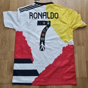 Unik Ronaldo fotbollströja med flera klubbmärken - Pris kan diskuteras. Säljer en färgstark och unik kortärmad fotbollströja med Ronaldo 7 på ryggen. Tröjan har klubbmärken från Real Madrid, Juventus, Manchester United, Sporting Lissabon och fler. Färgerna är vit, gul, röd och svart med tryckta detaljer och loggor.