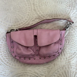 Rosa axelväska med nitar från Unlimited - Säljer en rosa axelväska från Unlimited i skinn med två stora fickor framtill och dekorativa silverfärgade nitar längs kanterna och axelremmen. Väskan har dragkedja upptill och ett innerfack med dragkedja. Perfekt för dig som vill sticka ut med en cool accessoar.