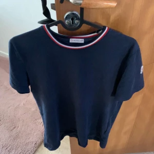Mörkblå t-shirt från Moncler - Säljer en mörkblå t-shirt från Moncler med rund halsringning som har röd och vit kant. Klassisk och stilren design med Moncler-logga på ärmen. Perfekt för en clean och snygg look. Priset kan diskuteras 