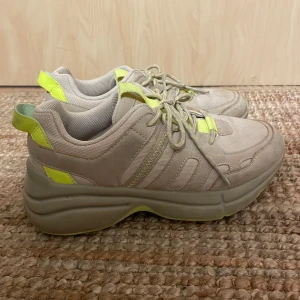 Beige sneakers med neongula detaljer - Chunky sneakers i beige med neongula detaljer på häl, plös och sidor. Skorna har snörning och en rejäl plattformssula som ger extra stöd. Väldigt bra skick, ingen doft.