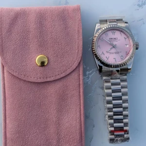 Seiko klocka med rosa urtavla och stållänk - Stilren Seiko klocka med ljusrosa urtavla, datumvisning och romerska siffror. Boett och länk i silverfärgat stål. Automatisk rörelse synlig genom glasbaksida. Levereras med rosa fodral i mockaliknande material.