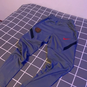 Grå Nike PSG träningsbyxor Dri-Fit - Säljer ett par grå träningsbyxor från Nike med PSG-logga och röd swoosh. Byxorna är i Dri-Fit material, har resår i midjan och smal passform. Perfekta för fotboll eller träning. Snygga detaljer med svarta inslag vid knäna.