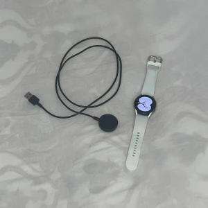 Vit Galaxy Watch4 med vit silikonarmband - Säljer en vit smartklocka från Samsung med runt ur och silikonarmband. Klockan har en slät design och kommer med en trådlös laddare. Perfekt för dig som gillar teknik och vill ha koll på hälsan. Den har Wifi Bluetooth GPS och LTE.