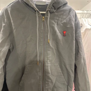 Grå hoodie med dragkedja från Ami - Grå hoodie med dragkedja från Ami, prydd med det klassiska röda hjärtlogotypen på bröstet. Jackan har huva med dragsko och två fickor framtill. Bra skick, nypris runt 3500-3800!