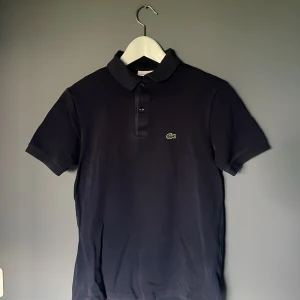 Marinblå pikétröja från Lacoste - Säljer en klassisk mörkblå pikétröja från Lacoste med korta ärmar och krage. Tröjan har två knappar framtill och den ikoniska gröna krokodilloggan broderad på bröstet. Perfekt för en stilren och sportig look. Inga defekter eller tecken på slitage. Skick 9/10💯