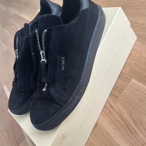 Svarta sneakers från Axel Arigato med dragkedja - Säljer ett par svarta sneakers från Axel Arigato i mocka med coola dragkedjor framtill istället för klassisk snörning. Skorna har används ett tag men de har inga stora skador. Skorna är lite smutsiga på bilden men dem är mycket bättre i verkligheten. Skorna är storlek 42.
