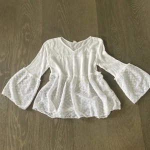 Vit blus med broderade detaljer och volangärmar - Säljer en söt vit blus med v-ringning och vida volangärmar. Nedre delen av blusen och ärmarna har broderade spetsdetaljer som ger en romantisk känsla. Perfekt att styla med jeans eller kjol för en fräsch look.