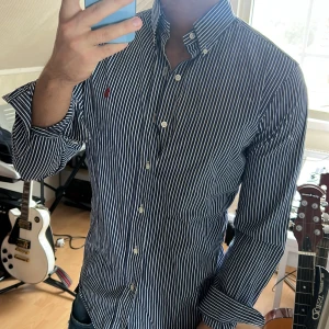 Blåvit randig skjorta från Polo Ralph Lauren - Säljer en blå och vit randig skjorta från Polo Ralph Lauren, custom fit. Skjortan har klassisk krage, knappar framtill och lång ärm. Ikoniska röda broderade loggan på bröstet. Perfekt för en stilren look.